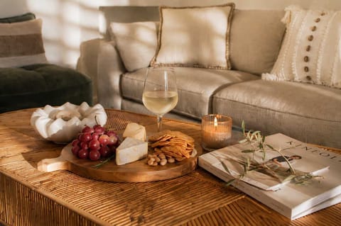 Settle in and relax in this inviting living room setting, featuring a woven rattan coffee table topped with a gourmet charcuterie board, a glass of white wine, a glowing candle, and soft plush sofa seating.