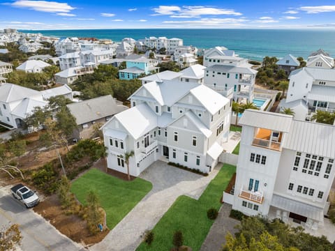 Aerial view of a spacious coastal retreat set in a charming beach community. The property features modern architecture, lush lawn, and driveway parking, surrounded by tropical landscaping and neighboring homes. The turquoise Gulf waters shimmer just beyond