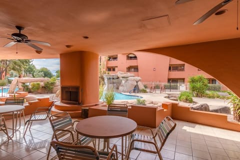 Covered patio features round tables, metal chairs, ceiling fans, and a built-in fireplace overlooking the pool area.