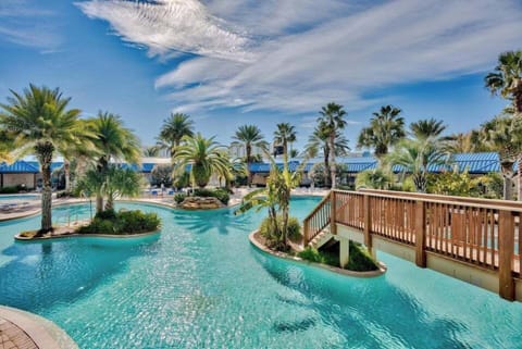 Resort-style pool w/ winding waterways, palm trees, and a sunny bridge connecting lush islands.