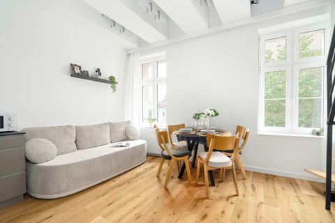 A bright living area combines a small sofa, dining table, and decorative shelving beneath white ceiling beams. Large windows bring natural light into the room.