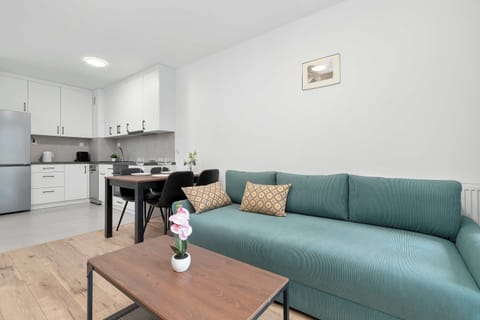 A modern open-plan living room with a teal sofa and a compact dining area. The space connects seamlessly to a bright kitchen, creating a functional and cozy layout.