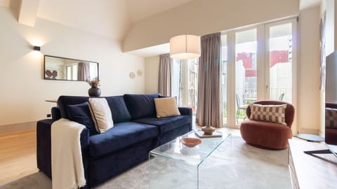 Bright and stylish living room with a plush navy sofa, elegant glass coffee table, and big windows opening onto the balcony. #LovelyStay #Airbnb