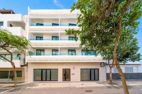 A modern white apartment building with blue glass balconies on a sunny tree-lined street.
