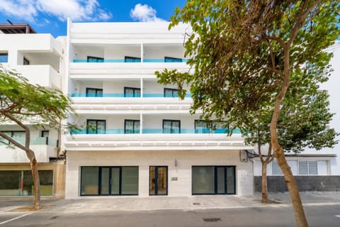 A modern white apartment building with blue glass balconies on a sunny tree-lined street.