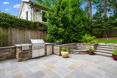 A secluded backyard sanctuary featuring beautiful garden landscaping, a huge stone terrace, and a premium outdoor kitchen with a natural gas grill and secondary two-burner stove built for entertaining.