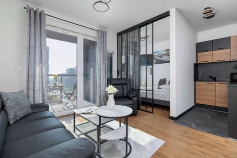 A modern living room features a dark sofa, a round glass coffee table, and a glimpse of a black and wood-toned kitchen area. Large windows or a glass door lead out to a balcony in the background.