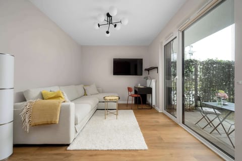 A bright, modern living room features a large cream-colored sofa with a yellow throw blanket resting on a fluffy white rug. Large floor-to-ceiling glass doors open directly onto a private, fenced-in garden patio.