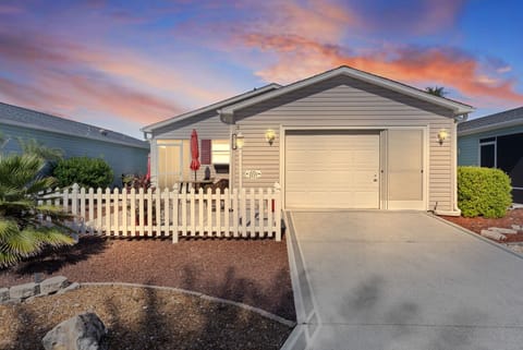 Charming single-story home with white picket fence, welcoming front porch, and stunning sunset backdrop painting the sky in vibrant coral and blue hues.