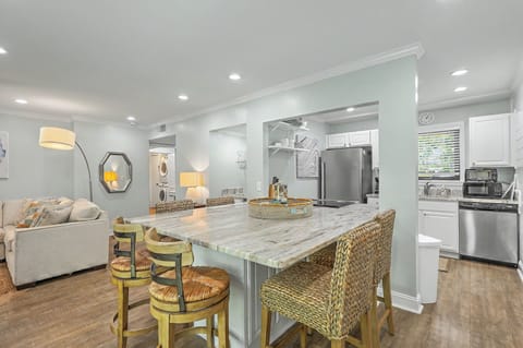 An open-concept kitchen features bright countertops and woven barstool seating at a spacious island. Stainless steel appliances and white cabinetry complement the modern coastal layout, offering a direct view into the living space.