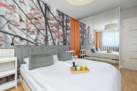 A modern bedroom features a large double bed dressed in white linens, with a small tray of fruit resting on top. A striking accent wall with a tree-patterned mural adds a bold touch of orange and grey to the space.