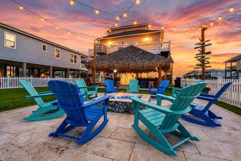 Twilight fire pit patio with Adirondack chairs, string lights, tiki pavilion, fenced yard and illuminated raised beach house.