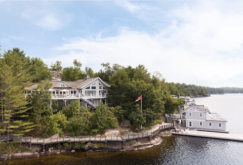 Lakefront cottage with gray siding, large windows, wraparound deck, and boathouse on rocky shoreline.