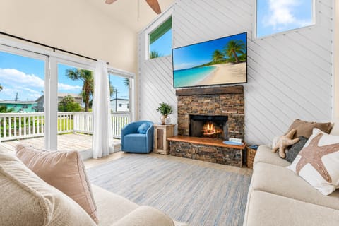 Bright living room with soaring ceilings, stone fireplace, large wall-mounted TV, and oversized windows bringing in abundant natural light from multiple directions.