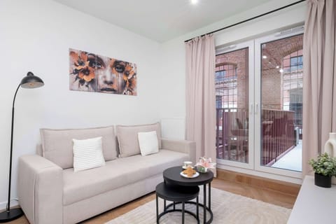 A cozy living room features a light beige sofa, a black coffee table, and a tall floor lamp in the corner. Large glass doors open up directly to an outdoor terrace area.