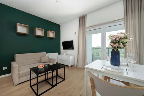 A modern living room features a dark green accent wall, a light beige sofa, and a black coffee table. To the right, a white dining table with chairs is visible near a large window.