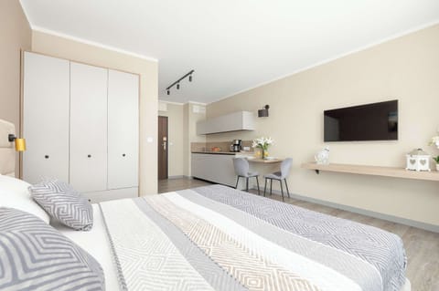 A modern studio apartment layout featuring a large double bed with gray and white patterned bedding. The background shows a white wardrobe and a minimalist kitchenette stretching along the wall.