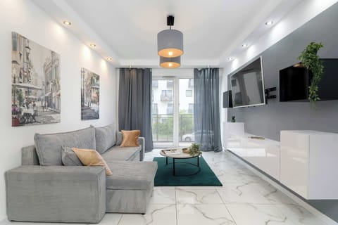 A modern living room features a comfortable grey sofa, a sleek white media konsola under a wall-mounted TV, and large windows that let in abundant natural light.