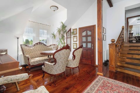 An elegant sitting area with patterned armchairs, a dining table, and polished wooden floors beneath exposed ceiling beams. Warm wood furnishings give the room a classic, traditional character.