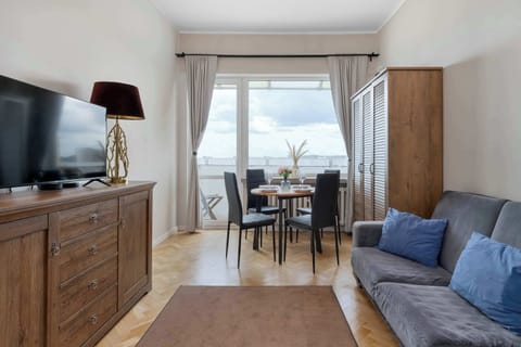A cozy living room features a grey sofa with blue accent pillows, a large wooden dresser with a TV on top, and a dining table set near a bright window in the background.