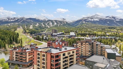 A mountain resort town nestled in a stunning alpine valley, surrounded by snow-capped peaks and evergreen forests, with ski slopes and nearby properties visible across the landscape.