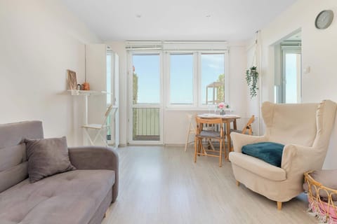 A bright and cozy studio apartment living area featuring a grey sofa, a comfortable armchair, and large windows leading to a balcony. Light wooden flooring and neutral walls create a spacious, inviting atmosphere.