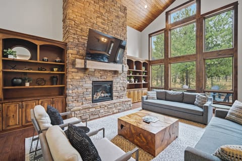 Relax in your sunlit living room with soaring ceilings, a stone fireplace, and floor-to-ceiling windows framing lush forest views—your perfect mountain retreat.