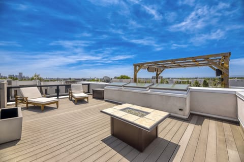 A clean, modern rooftop setup with light wood decking, two cushioned loungers, and a square fire pit under a simple wooden pergola. Glass railings open to a wide city skyline and blue sky, giving the whole space a calm, private‑escape vibe.