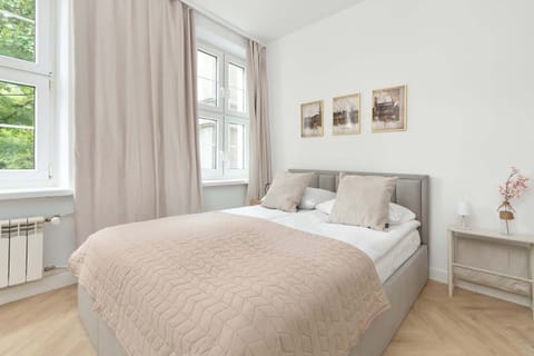 A bright bedroom area with a double bed, soft beige textiles, and large windows dressed with light curtains. Minimalist décor and framed artwork create a calm atmosphere.