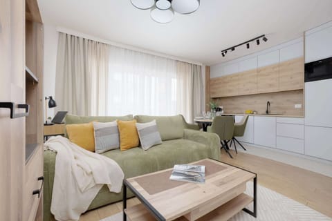 A bright living space showcases a light green fabric sectional sofa accented with yellow and gray patterned pillows. A low wooden coffee table with a metal frame sits in the center of the room on light wood flooring.