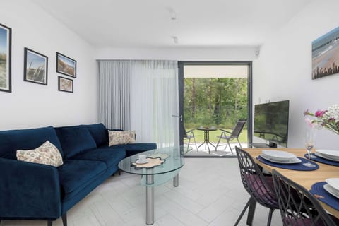 A bright living area features a deep blue sofa adorned with patterned pillows, situated next to a glass coffee table. Large floor-to-ceiling windows open directly onto an outdoor terrace surrounded by lush green trees.