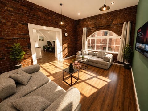 Spacious living room with exposed brick walls, wooden flooring, and two comfortable grey sofas. Large arched window brings in natural light, while ceiling lights brighten the space. Open layout connects directly to the dining and kitchen area.