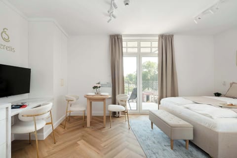 A spacious, modern living area featuring a plush double bed, a sleek TV console, and elegant light flooring. Large windows flood the space with natural light, creating a bright and welcoming atmosphere.