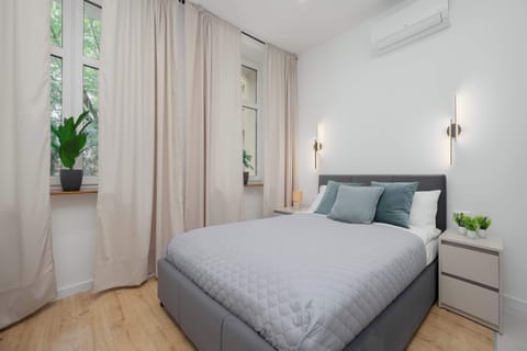 A compact studio apartment features a comfortable double bed dressed in light gray quilted linens. Large windows covered with neutral floor-to-ceiling drapes fill the room with soft daylight.