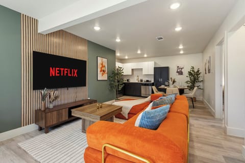 Bright open-plan living area with a bold orange sectional, large wall-mounted TV, warm wood accents, and easy flow to the kitchen and dining space for relaxed time together.