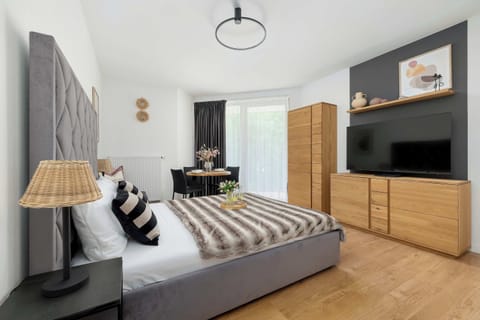 The view of the bedroom from another angle reveals a wooden chest of drawers placed underneath a large, wall-mounted TV. Next to the bed with its quilted headboard sits a bedside table with a classic wicker lamp.