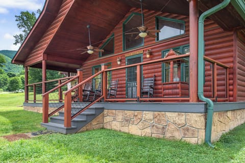 Charming log cabin exterior with a wraparound covered porch featuring rocking chairs, nestled among lush green lawns and mountain views—a peaceful retreat where rustic meets relaxation.