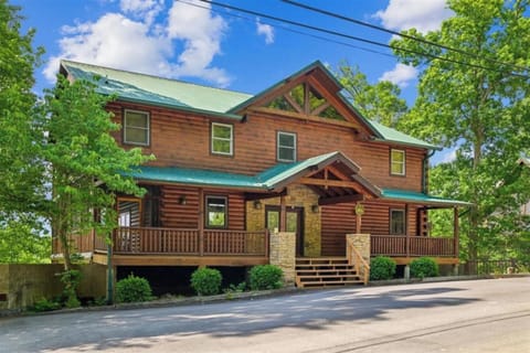 A spacious two-story log cabin with green metal roofing, stone accents, and wrap-around porches, nestled among mature trees in a mountain resort setting.
