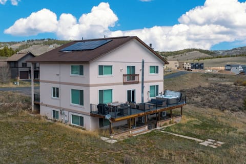Multi-level mountain retreat with expansive deck and hot tub, surrounded by rolling hills and nearby residential area under open sky.