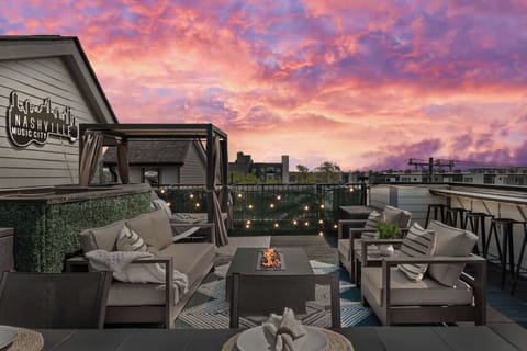 The rooftop terrace features a cozy fire pit lounge, plush cushioned outdoor seating, a draped pergola, and warm string lights lining the ivy privacy wall and breathtaking sunset skyline views overhead. - The Wild Rose