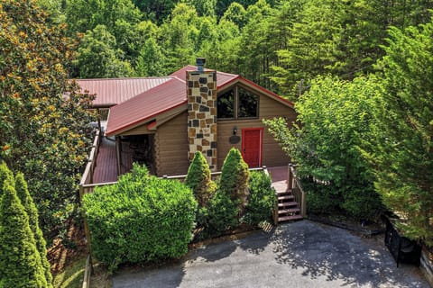 A charming mountain cabin with a distinctive stone chimney and red metal roof nestled in lush forest. The welcoming red door and well-maintained landscaping create an inviting entrance to this secluded retreat.