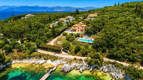 An aerial drone photograph of a peach-colored luxury villa with a private pool, completely surrounded by dense green forest on a rocky coastline with clear turquoise water.