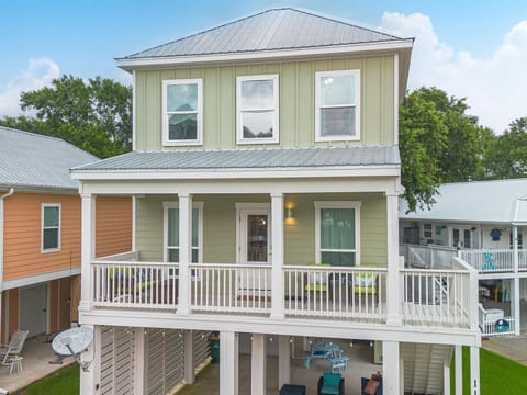 Elevated beach house with green board-and-batten siding, white wraparound porch railing, and a covered lower entertainment area
