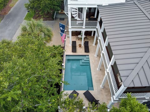Aerial view of a charming vacation rental featuring a private pool, spacious patio with loungers, multi-level balconies, and lush landscaping along a quiet residential street.