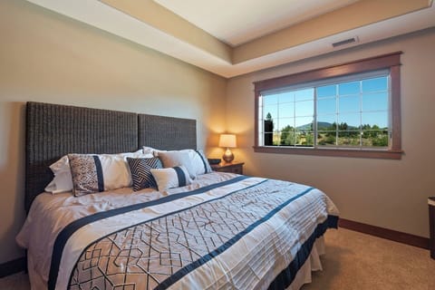 A modern bedroom setting featuring a king-sized bed with a high woven dark headboard, geometric patterned bedding in white and navy, a small wooden nightstand with a lamp, and a large window showcasing a green hillside view.