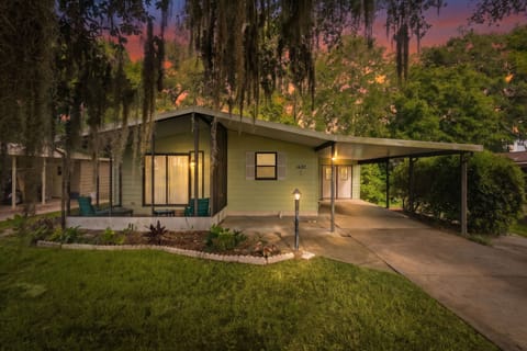This charming mid-century modern retreat is framed by majestic trees draped in Spanish moss, with a vibrant sunset painting the sky in shades of pink and orange.