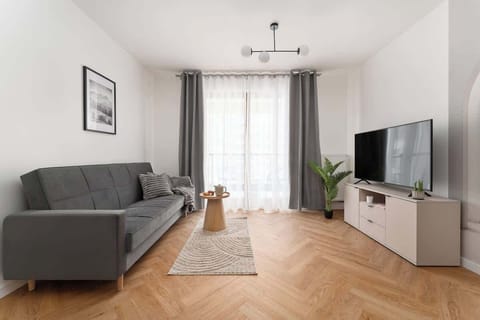Salon wyposażono w wygodną, rozkładaną sofę, utrzymaną w stonowanych, nowoczesnych odcieniach szarości. Naprzeciwko znajduje się duży telewizor Smart TV z dostępem do kanałów telewizyjnych.
