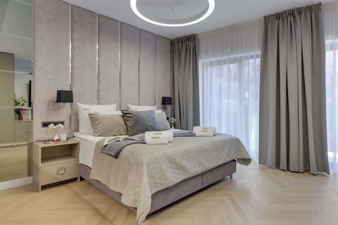 Otwarta przestrzeń z łóżkiem małżeńskim, Smart TV i miejscem do pracy. Wnętrze uzupełniają eleganckie detale, nastrojowe oświetlenie oraz pojemne szafy w przedpokoju.