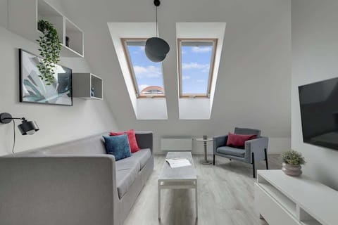 Przestronny, klimatyzowany salon urządzono w stonowanych barwach szarości i bieli, z delikatnymi akcentami czerni i zieleni. Goście mogą zrelaksować się na wygodnej, rozkładanej sofie lub fotelu, oglądając ulubione programy na telewizorze. 