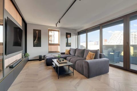 Salon utrzymany jest w nowoczesnej, minimalistycznej stylistyce, w której dominują ciemne kolory, naturalne drewno oraz oświetlenie LED. Znajduje się tu duża, rozkładana sofa, a naprzeciw niej zamontowano telewizor z kanałami TV oraz Smart TV.